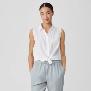 A woman with her hair pulled back wears a white, sleeveless linen blouse with a knot tied in front of the stomach and grey linen pants. She has her hands tucked in the pockets.
