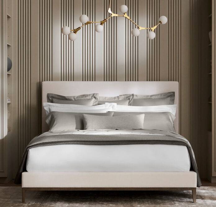 A white platform bed with crisp white sheets and duvet, accented by steel grey pillows and coverlet, sits in a modern room against a back drop of white and black striped wallpaper.