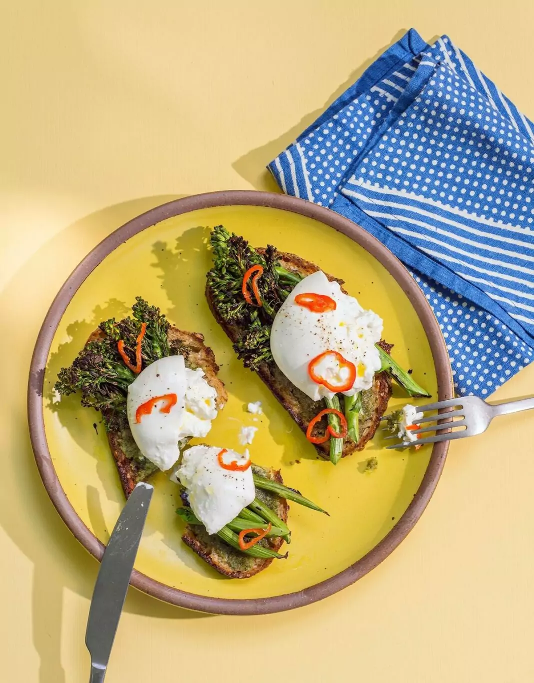 Yellow plate with two pieces of toast. On top of the toast is burrata cheese and some charred pieces of Broccolini sprinkled with sliced red pepper.