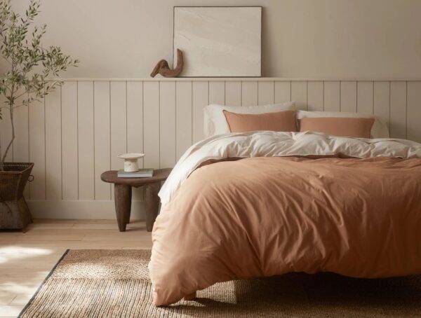 A bed with clay-colored Coyuchi bedspread and pillows and cream-colored sheets in a room with a minimalist room with a potted tree, small wooden side table, and shiplap panelling on the walls.