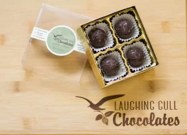 Four chocolate caramel truffles from Laughing Gull Chocolate sit in a gold box on a bamboo board.