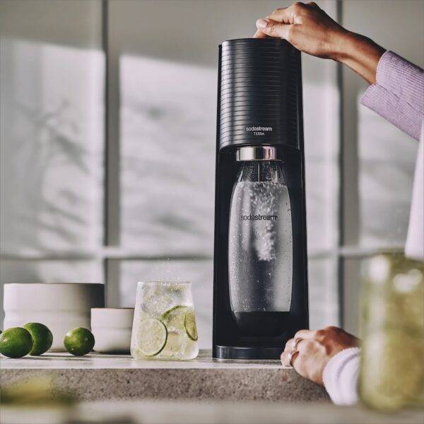 In a brightly lit room, on a kitchen counter, a woman's hand pushes the button on top of a black SodaStream machine, which turns flat water into sparkling water. A glass of very bubbly water contains slices of fresh lime.