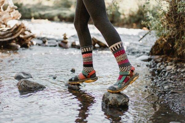 A woman's legs, shown in black leggings and colorful, mismatched socks and sandals, crosses a small stream on tiptoe over small rocks.