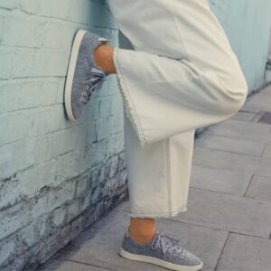 We see the legs of a woman wearing wide-legged white pants and grey wool tennis shoes made by Allbirds. She leans against the side of a building with light blue brick.