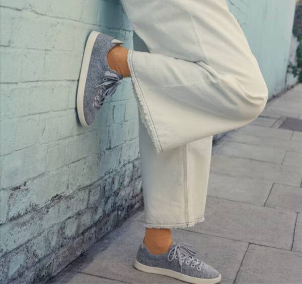 We see the legs of a woman wearing wide-legged white pants and grey wool tennis shoes made by Allbirds. She leans against the side of a building with light blue brick.