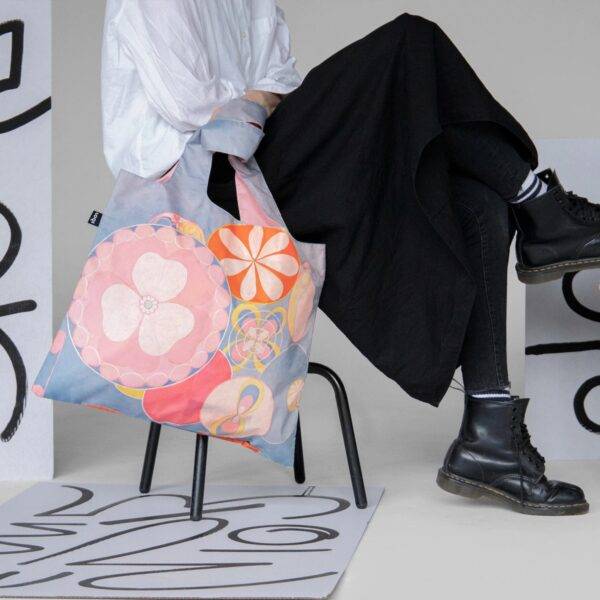 From the neck-down, we see a person wearing black pants and shoes and a white shirt holding a Loqi reusable bag featuring a print by artist Hilma of Klint. The bag has a pattern of greys, pinks, reds, and yellows. The person sits on a chair in a white room painted with black squiggles.