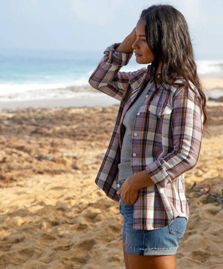 A woman stands on a beach in cutoff jean shorts and a mauve and cream colored flannel shirt from sustainable clothing brand Outerknown. Photo Credit Outerknown.