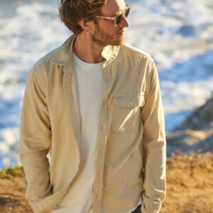 A man wearing sunglasses, a beige corduroy shirt, and white t-shirt, stands on a cliff above a beach.
