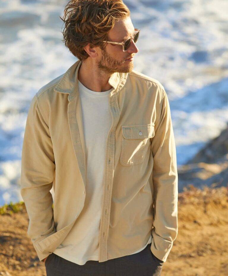 A man wearing sunglasses, a beige corduroy shirt, and white t-shirt, stands on a cliff above a beach.