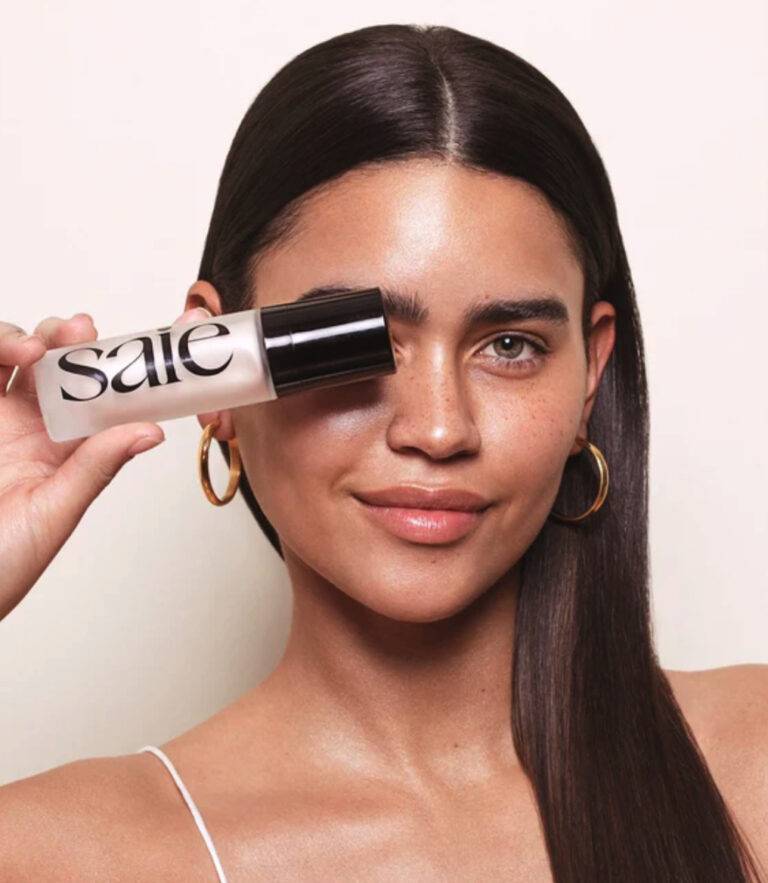 A woman with radiant, freckled, olive-toned skin, dark hair and thick dark eyebrows holds up a package of Saie Beauty's Glowy Super Gel in Starglow. Photo Credit - Saie Beauty