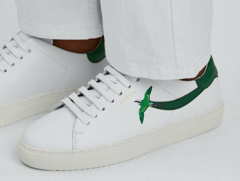 A person from the ankles down, shown in white jeans and a pair of white tennis shoes from designer Axel Arigato, with a green stripe on the side of the shoe that turns into a green birds with wings spread.