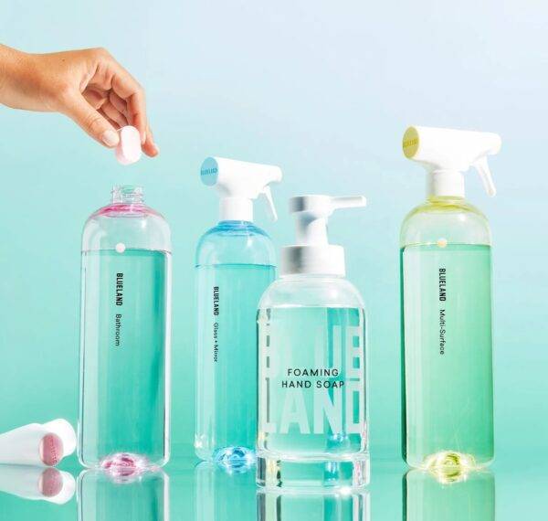 A turquoise background shows four bottles of cleaning solution made by Blueland. A woman's hand hovers over one of the bottles to drop a tab into the bottle.
