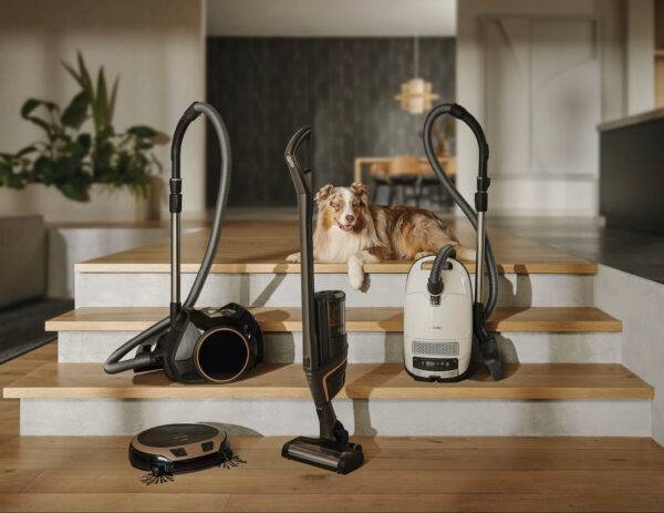 Two canister vacuums, a robot vacuum, and a cordless vacuum are displayed on a staircase. An Australian Shepherd dog sits at the top of the stairs. Photo Credit - Miele