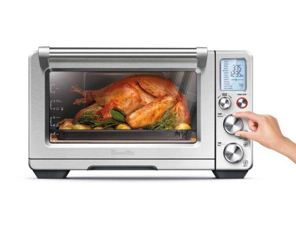 A large countertop smart oven made by Breville with a roast turkey inside of it, with a woman's hand adjusting one of the knobs.