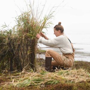 A woman works in front of a body of water, sitting on top of hay wearing boots and a pair of brown overalls made by Dovetail Workwear.