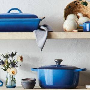 Two shelves of cookware hold an assortment of bright, Azure-blue enamel-coated cast iron cookware from Le Creuset, including a round Dutch oven and a covered bakeware dish, as well as a molcateje, a dish cloth, and some flowers and gourds.