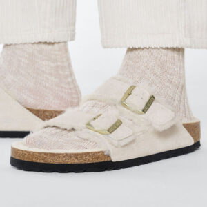 A person's feet shown in cream-colored suede, two-strap sandals with a white fuzzy lining, wearing oat-colored socks underneath and white pants. Photo credit: Birkenstock