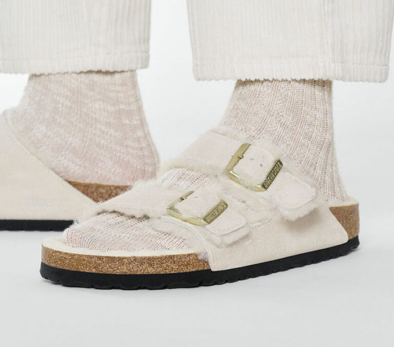 A person's feet shown in cream-colored suede, two-strap sandals with a white fuzzy lining, wearing oat-colored socks underneath and white pants. Photo credit: Birkenstock