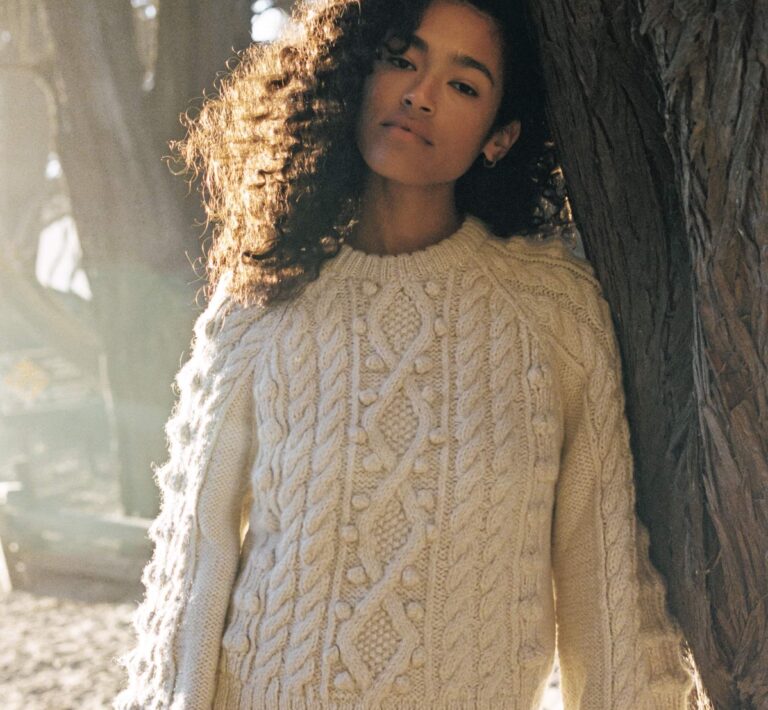 A woman wearing a thick woolen cream-colored sweater leans against a tree with sunlight streaming down behind her.
