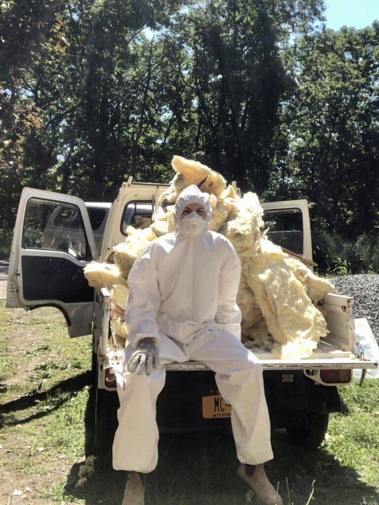 Bea sits on back of pickup truck full of insulation wearing a full-body white jumpsuit and mask.