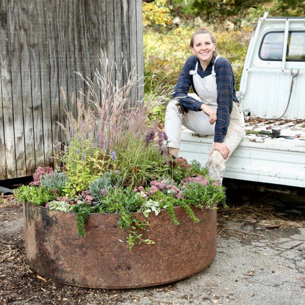 Bea sits on the back of her small white truck and smiles to camera with a large raised flower planter in the foreground.