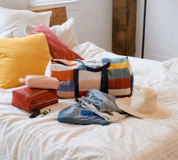 A bed made up in white linens and red and yellow pillows has items on it for packing, including a colorful striped duffel bag from brand Anchal, jeans, a hat, and a toiletry bag.