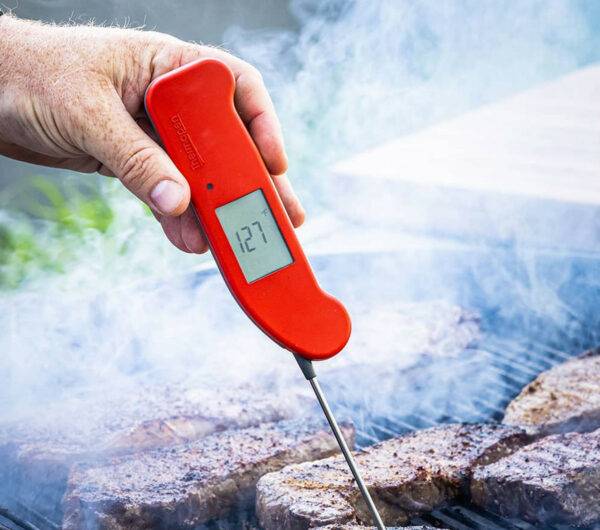 A hand holds a red Thermapen One thermometer pen over steaks on a grill to take the internal temperature of the meat.