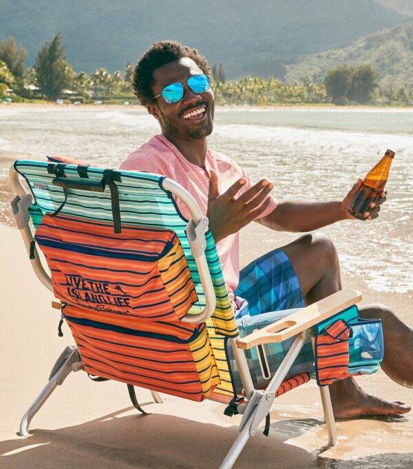 A smiling man in sunglasses sits on a beach in a colorful striped beach chair.