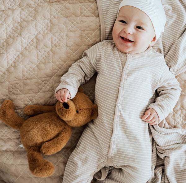 A baby in a beige onesie and hat lies on a blanket and smiled while touching a teddy bear.