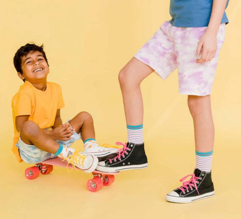 A smiling child in blue tie-dye shorts and a yellow shirt sits on a skateboard while a bigger kid in purple tie-dye shorts stands next to the skateboard.