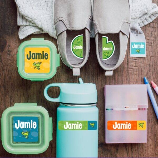 Two food containers, a pair of slip-on tennis shoes, a sweater, a box of crayons, and a water bottle all feature prominent colorful labels with frogs and the name Jamie.
