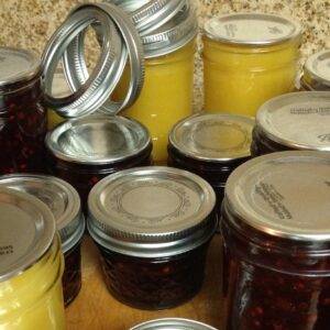 Assorted clear glass canning jars, some filled with creamy yellow curd and others with dark red jam, sit on a cutting board with the rings close by to screw on top of the lids.