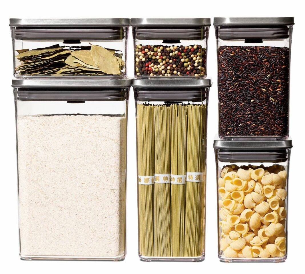 A set of six clear containers of varying sizes with stainless steel tops contain foods including bay leaves, whole wheat flour, peppercorns, noodles, rice, and pasta shells.