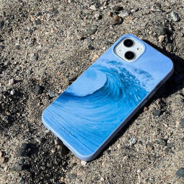 A cell phone screen-side down in a blue case showing a wave design and the brand name "pela" sits on sandy, rocky ground.