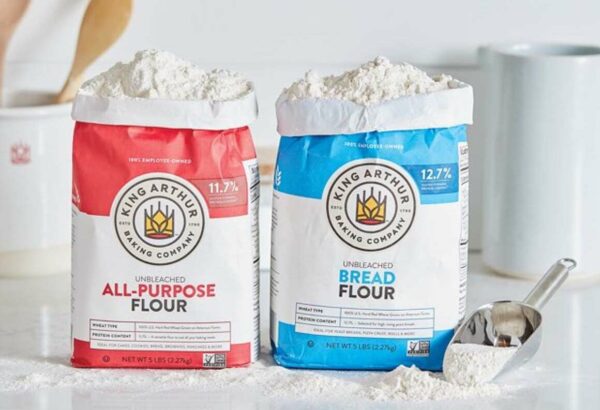 Two open, overflowing bags of flour from King Arthur Baking Company on a white kitchen counter top, with flour scattered next to them.