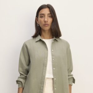 A brunette woman wears a sage green linen blouse, unbuttoned, over a white tee-shirt and cream-colored pants.
