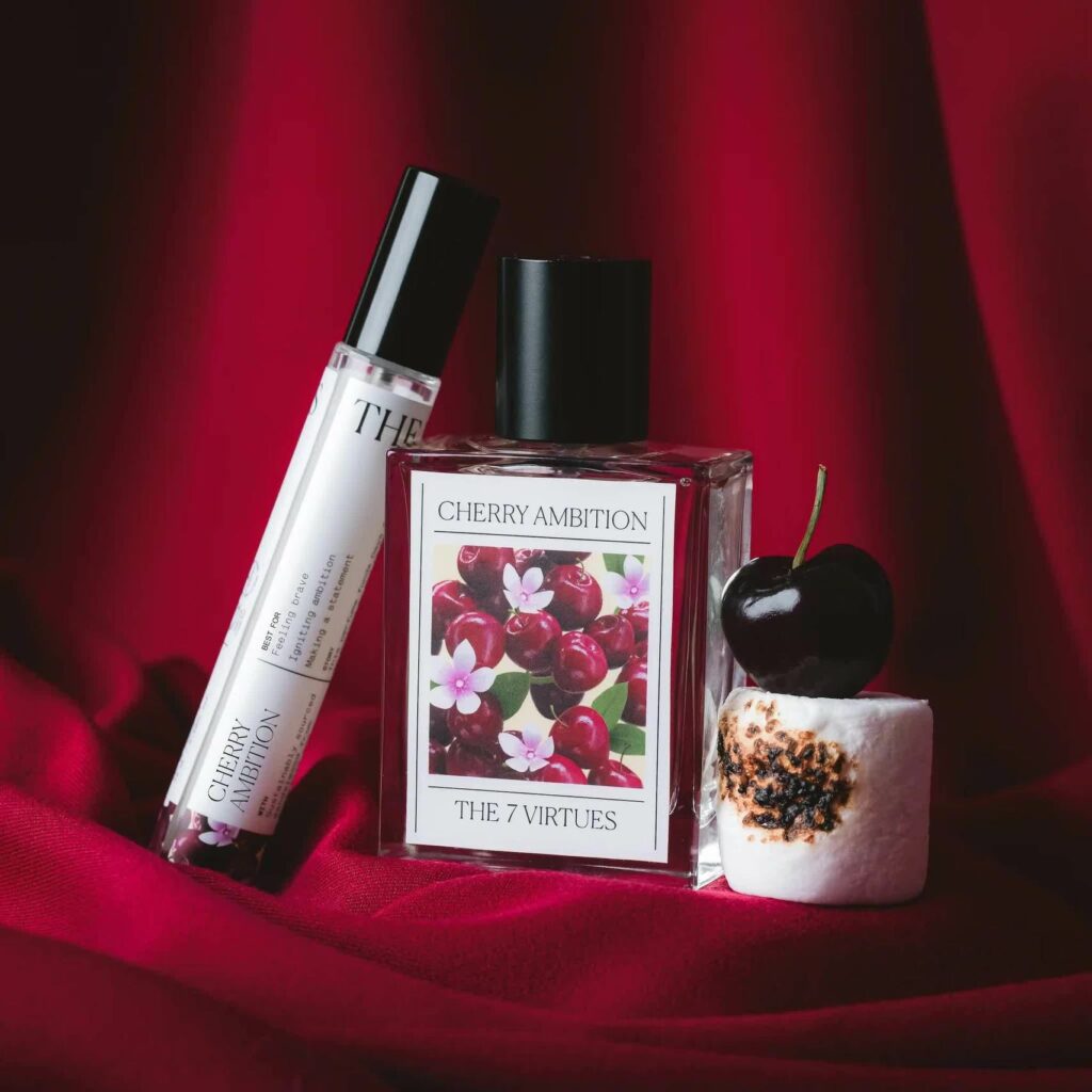 A bottle of perfume that reads with a red cherry and pink-flower-adorned label that reads Cherry Ambition / The 7 Virtues sits on a piece of red satin, with a travel sized bottle of the perfume on one side of it and a toasted marshmallow with a fresh red cherry on the other side.