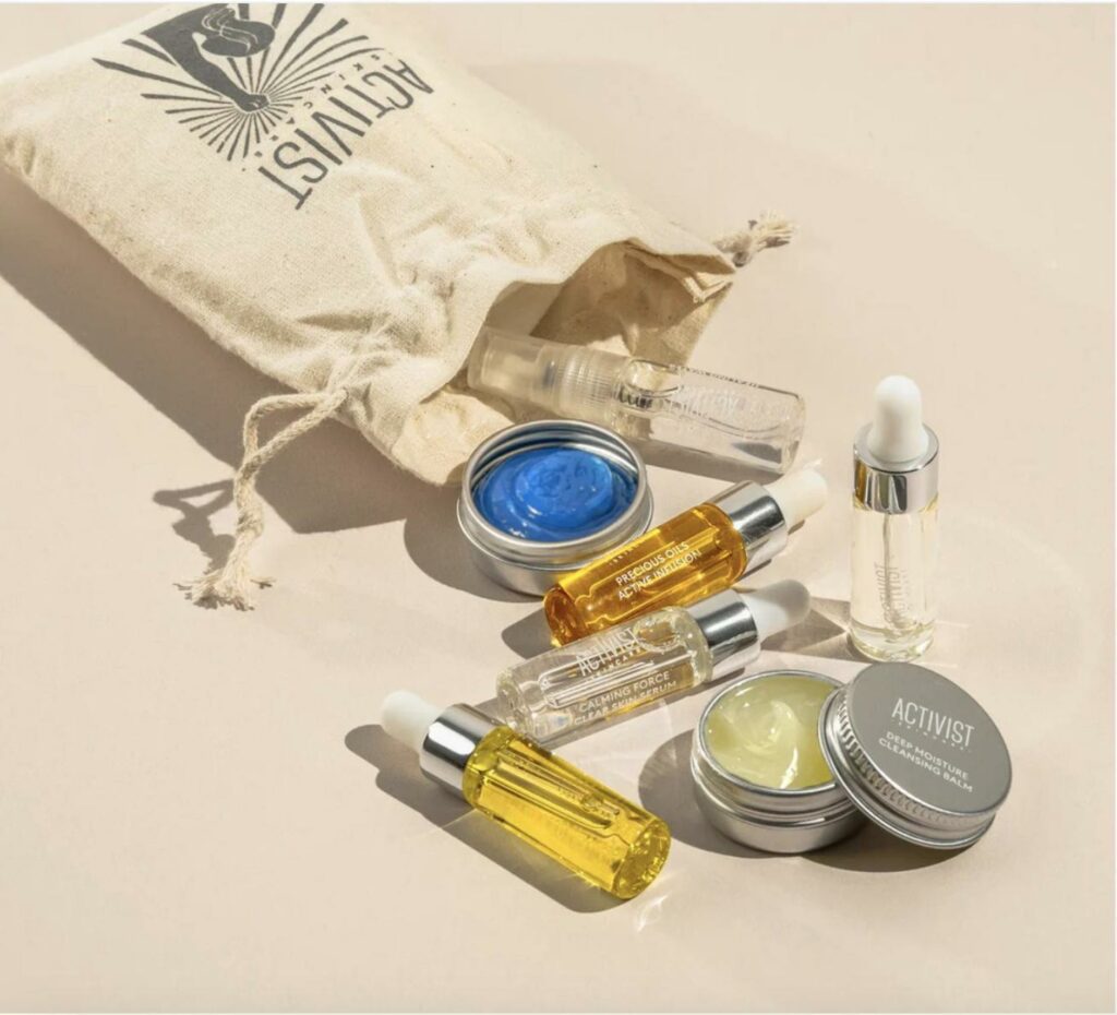 A small burlap bag with the word Activist on it is open on its side with small assorted vials and tins of skincare products. 
