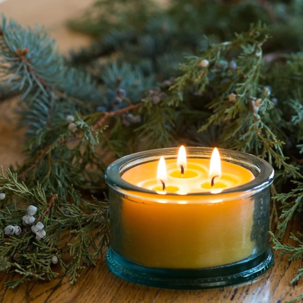 A beeswax candle in a clear glass vessel, shown with pine tree boughs