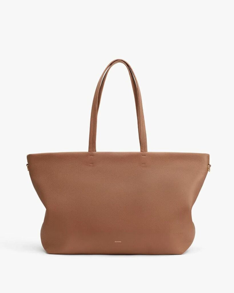 A large caramel-colored leather tote bag with thin straps and small, foil-stamped branding reading Cuyana on the bottom.