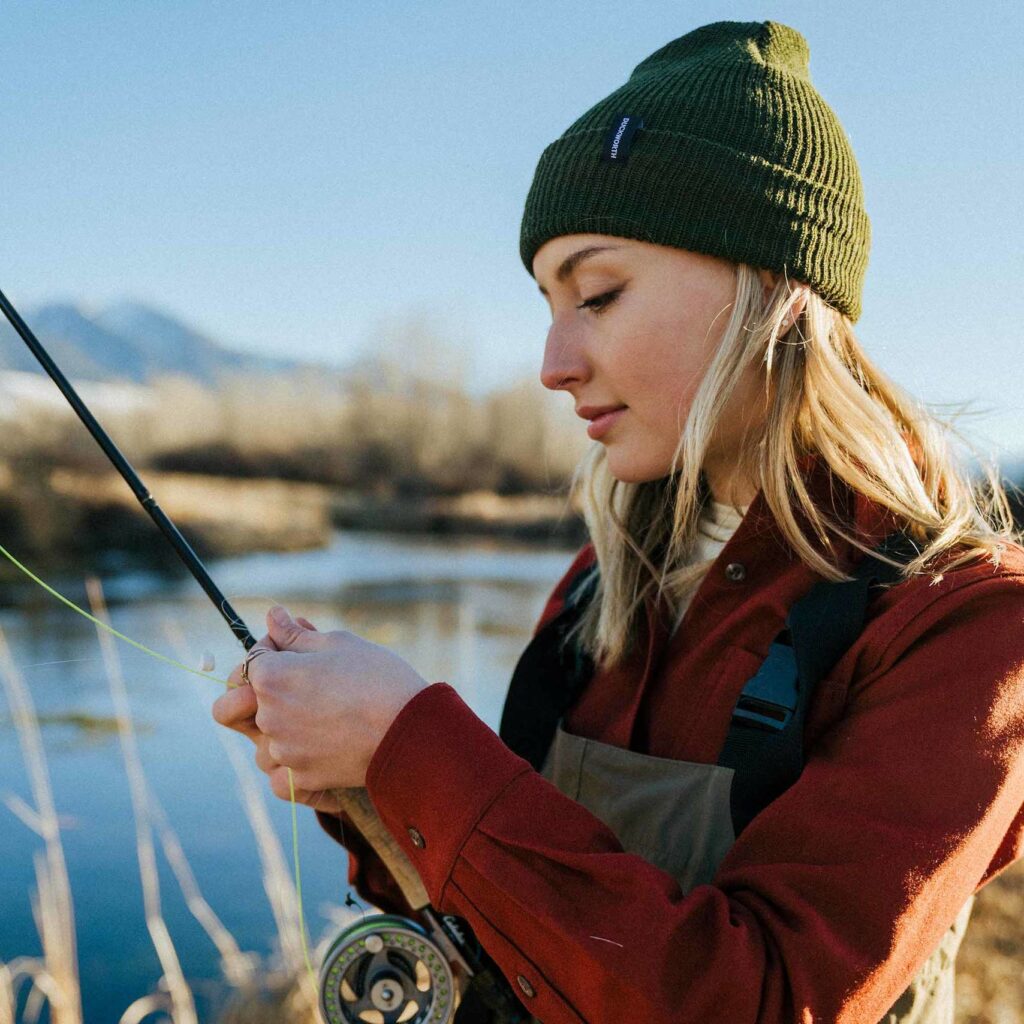 A woman wearing fishing gear and an olive green beanie untangles fishing line from her fishing rod.