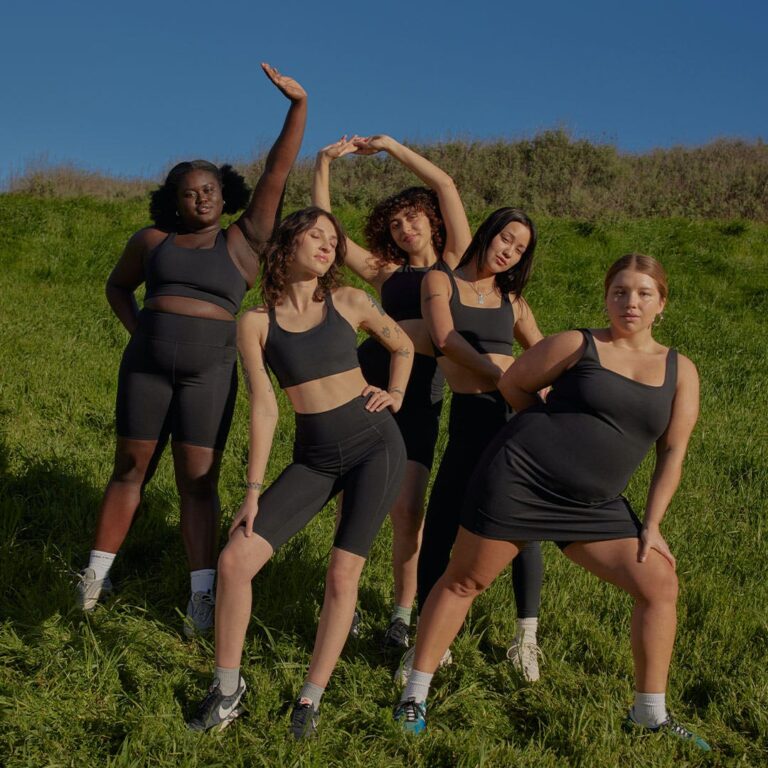 Five women of varying ethnicities and sizes stand on a grassy hill and wear similar sustainable workout clothes in brown.
