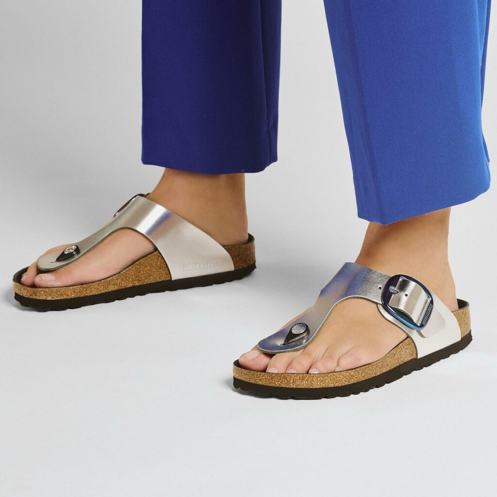 From the mid-thigh down, a woman’s legs shown in blue slacks and a pair of metallic silver Birkenstock sandals with a single thong toe strap. 