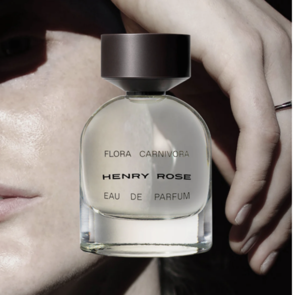 A close-up on a woman’s face and hand with a clear perfume bottle in front of it that reads Henry Rose Flora Carnivora Eau de Parfum. 