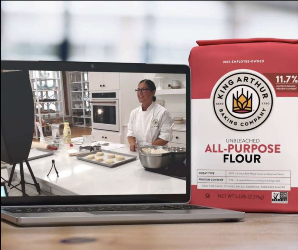 A laptop is open on a wooden counter and the screen shows a chef in front of a baking tray of scones; a red and white bag of King Arthur Unbleached All-Purpose Flour is on the counter next to the laptop