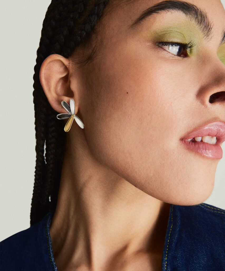 A close-up on the face of a woman in lime-green eyeshadow wearing an earring that looks like a deconstructed flower petal with four silver petals and one gold petal.
