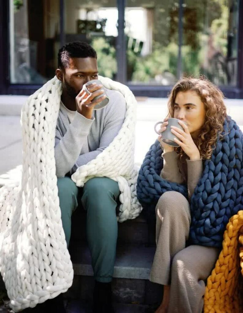 A young man and a young woman drink coffee outside, each with a chunky knit blanket draped over their laps.