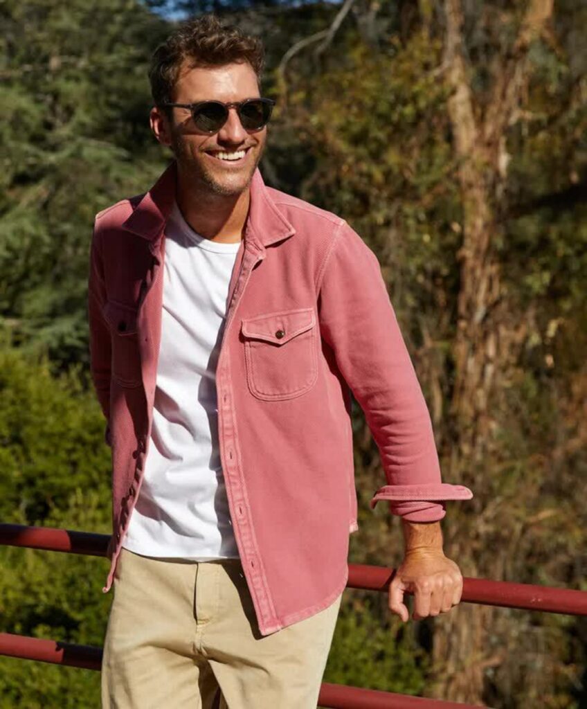 A man in sunglasses stands against a railing in khakis and a faded red button-up shirt.