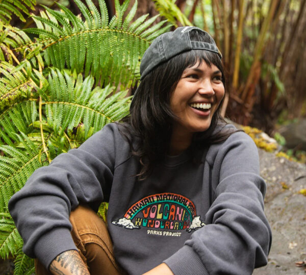 A smiling woman sits on a wet rock surrounded by fields wearing a backwards baseball cap and a faded black sweatshirt with a rainbow patch embroidered on it that reads HAWAII NATIONAL PARK / VOLCANIC WONDERLAND / PARKS PROJECT.