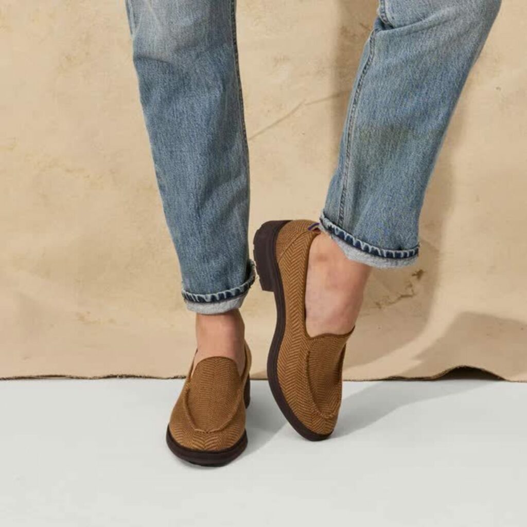 From just above the knees down, a person’s legs shown in medium-wash denim jeans rolled at the ankles and a pair of brown loafers with low black heels.